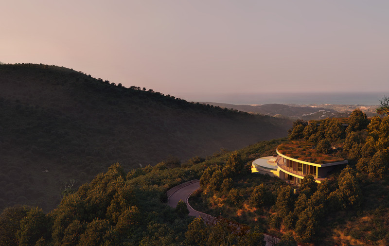 Villa I3 – Vitae Villas – Benahavís - aerial exterior view of curved green-roofed villa on a forested hillside at dusk with coastal views
