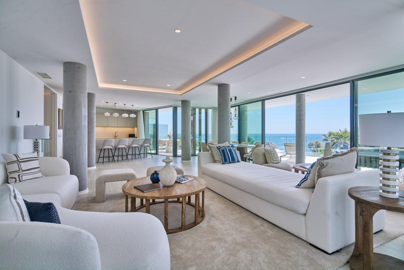 The Sapphire – Estepona - open-plan living room with white sofas, concrete columns, and panoramic sea views through floor-to-ceiling glass